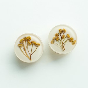 以下が含まれることがあります： 黄色いドライフラワーが閉じ込められた、丸い白いレジン製のイヤリングが2つ