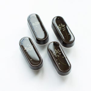 Könnte beinhalten: Vier dunkelbraune, rechteckige Schmuckschalen mit abgerundeten Kanten. Jede Schale zeigt ein zartes Blumendesign, das in klarem, glänzendem Harz eingeschlossen ist. Die Schalen sind auf einer weißen Oberfläche angeordnet und erzeugen eine saubere und elegante Ästhetik.