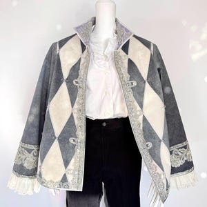 Könnte beinhalten: Eine Jacke im Harlekin-Stil mit einem rautenförmigen Muster in Grau und Creme. Die Jacke hat einen verzierten silbernen Besatz, dekorative Knöpfe und einen lila Kragen. Die Ärmel haben weiße Spitzenbündchen. Die Jacke wird auf einer Schaufensterpuppe präsentiert.