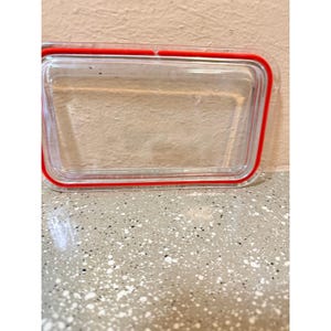 Puede incluir: Tapa transparente y rectangular para recipientes de alimentos, con un sello de silicona roja. La tapa está hecha de plástico transparente y está diseñada para adaptarse a un recipiente rectangular. El sello rojo proporciona un cierre hermético.