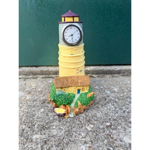 May include: A lighthouse-shaped desk clock featuring a yellow tower with a purple roof and a circular analog clock face with black hands. At the base sits a small yellow cottage with a brown thatched roof, green grass, and rocky terrain.