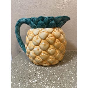 May include: A ceramic pitcher shaped like a pineapple. The pitcher has a yellow and cream-colored body with a textured surface. The handle and top are green, mimicking the pineapple's leaves. The pitcher is designed for serving beverages.
