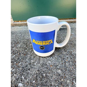 Könnte beinhalten: Weiße Keramiktasse mit einem blauen Feld mit dem Text "GOOD MORNING AMERICA" in Gelb und dem ABC-Logo. Die Tasse hat einen gebogenen Henkel und steht auf einer strukturierten grauen Oberfläche.
