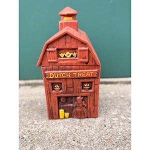Könnte beinhalten: Ein brauner Keramikbehälter in Scheunenform mit den Worten "DUTCH TREAT" in gelben Buchstaben. Der Behälter hat einen kleinen Turm und dekorative Elemente, darunter eine Figur in niederländischer Tracht.