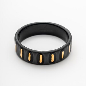 Puede incluir: Un anillo negro con detalles verticales dorados. El anillo es circular y está sobre un fondo blanco. La palabra "CARPESITA" está escrita en cursiva en la parte inferior de la imagen.
