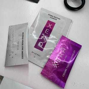 May include: Assortment of skincare product packets in silver, white, and purple. The packets include text such as "Exosome Daily Repair Hydrator", "Dermal Prepping Cleanser", and "Exosome Tightening Repair Lift". A black ring and a dark packet are also visible.