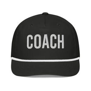 May include: Black baseball cap with the word "COACH" embroidered in white block letters. The cap has a white rope detail along the brim.