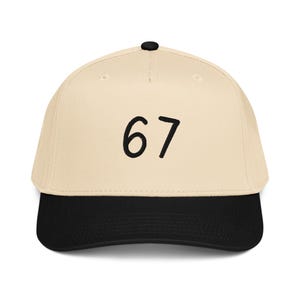 May include: A baseball cap with a cream-colored crown and a black brim. The number "67" is embroidered in black on the front of the cap. The cap has a curved brim and a black button on top.