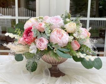 May include: A floral arrangement featuring pink and peach peonies, eucalyptus, and other greenery. The flowers are arranged in a bronze-colored, footed urn. The arrangement is set on a white surface with a sheer fabric. The Floral Artisans Studio is printed on the bottom right.