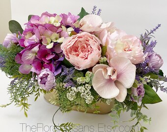 May include: A floral arrangement featuring pink peonies, purple hydrangeas, and lavender sprigs in a gold-colored oval container. The arrangement includes green foliage and small white flowers, creating a vibrant and colorful display. TheFloralArtisans.Etsy.com is visible.