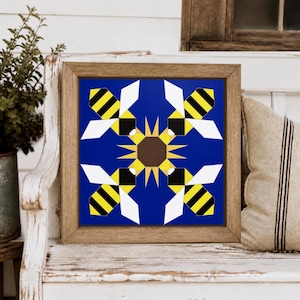 May include: A square framed wall art piece featuring a vibrant royal blue background. The design displays four stylized bees with yellow and black striped bodies and white wings, arranged symmetrically around a central brown and yellow sunflower.