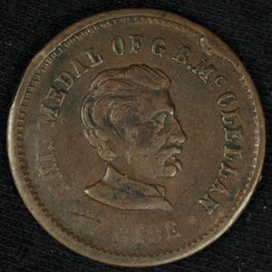 May include: A bronze-colored coin featuring a profile of a man with a mustache and the inscription "THE MEDAL OF G.B.M. MCCLELLAN". The coin has a detailed design and a worn appearance, suggesting age and historical significance.