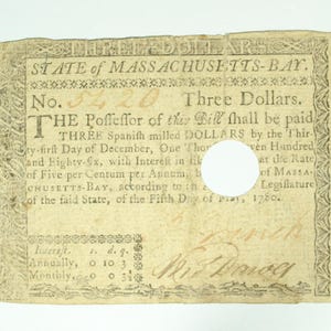 May include: An antique three-dollar bill from the State of Massachusetts-Bay, dated May 5, 1780. The aged paper currency features ornate borders, text, and a handwritten signature. The bill is torn with a white circle in the center.