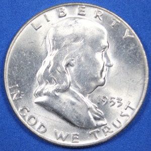 May include: A silver 1953 half dollar coin featuring a profile of Benjamin Franklin. The coin has the inscription "LIBERTY" above Franklin's head and "IN GOD WE TRUST" below. The date "1953" is visible on the right side of the coin.