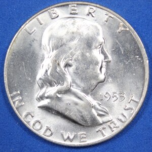 May include: A silver 1953 Franklin half dollar coin with the inscription "LIBERTY" above the portrait of Benjamin Franklin and "IN GOD WE TRUST" below. The date "1953" is visible on the right side of the coin.