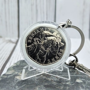 Könnte beinhalten: Ein silberner Schlüsselanhänger mit einer Münze, die Benjamin Bunny zeigt. Die Münze befindet sich in einem transparenten, runden Gehäuse. Der Schlüsselanhänger hat einen silbernen Ring und einen Karabiner.