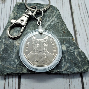 Può includere: Un portachiavi argentato con un porta monete in plastica trasparente che contiene una moneta d'argento. La moneta ha l'immagine di un gatto con le parole "Tom Kitten" attorno al bordo.