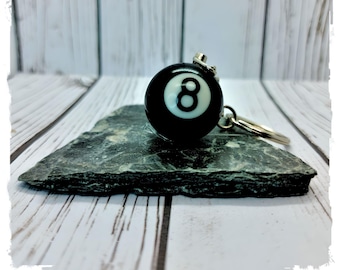 May include: Black and white 8 ball keychain with a silver ring and chain. The keychain is sitting on a dark gray slate surface.