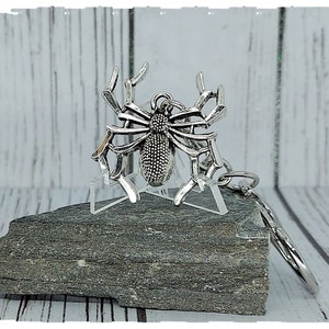May include: A silver spider keyring with a detailed design. The spider is sitting on a small, grey rock.