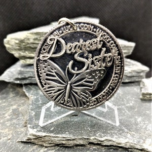 Puede incluir: Encanto de moneda de plata con un diseño de mariposa y las palabras "Dearest Sister" grabadas en el frente. La moneda está en un soporte negro.