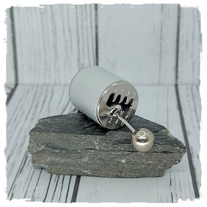 Op de afbeelding: Een zilveren versnellingspook-sleutelhanger met een witte cilinder en een ronde knop. De sleutelhanger heeft de nummers 1 tot en met 5 en de letters R en N.