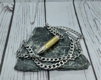 Può includere: Una collana a catena d'argento con un ciondolo a forma di proiettile. Il proiettile è in ottone e ha un colore dorato. La collana è appoggiata su una pietra grigia.