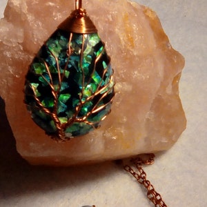 May include: A teardrop-shaped pendant necklace with a copper wire tree design. The pendant is made of iridescent green and blue glass pieces. The necklace is on a copper chain.