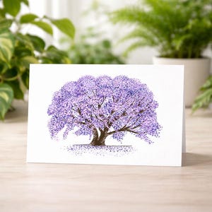 May include: A white greeting card with a watercolour illustration of a Jacaranda tree in full bloom. The tree has a brown trunk and branches, with a large, rounded canopy of purple flowers. The card is set on a light wood surface.