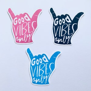 May include: Three stickers featuring a hand gesture design and the words "Good Vibes Only." The stickers are in pink, navy blue, and light blue, each with a distressed texture. The background is white.