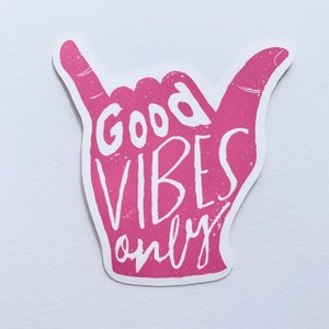 May include: A pink sticker in the shape of a hand making the shaka sign. The words "Good Vibes Only" are printed in white, with a distressed texture. The sticker is on a white background.