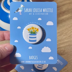 May include: A round button pin with a floral design featuring yellow daffodils in a blue and white striped pitcher. The pin is on a blue card with a seagull illustration and the text "SARAH LOUISA WHITTLE ART & ILLUSTRATION" and "BADGES".