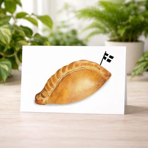 May include: A greeting card with a watercolour illustration of a Cornish pasty, golden-brown in colour, with a small black and white flag. The card is on a white surface, with green plants in the background.