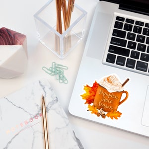 May include: A sticker featuring a brown mug with white whipped cream and a cinnamon stick. The mug is filled with a brown liquid and has the text "Pumpkin Spice Season" on it. The mug is surrounded by orange and yellow fall leaves and acorns.