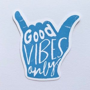 May include: A blue sticker in the shape of a hand gesture, with the words "Good Vibes Only" in white lettering. The sticker has a distressed, textured appearance, and is set against a white background.