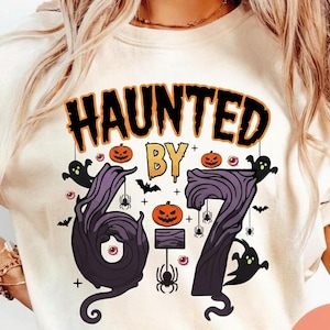 Może przedstawiać: Kremowa koszulka z grafiką o tematyce Halloween. Projekt zawiera napisy "HAUNTED BY 67" w kolorze czarnym i pomarańczowym, a także dynie, duchy, nietoperze i pająki. Cyfry 6 i 7 są stylizowane na ciemnofioletowy, drzewiasty wzór.