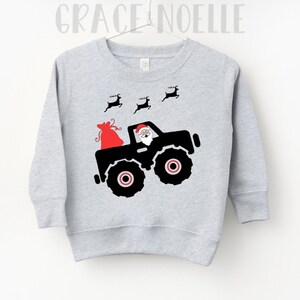 May include: A gray sweatshirt with a black silhouette of Santa Claus driving a monster truck with a red bag of gifts in the back. Three reindeer are flying above the truck.