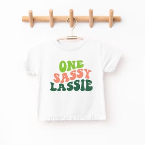 May include: White t-shirt with the text "ONE SASSY LASSIE" in green, pink, and white letters. The letters are in a retro font style.