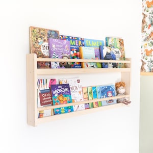 Peut inclure: Une étagère murale en bois clair à deux niveaux horizontaux. L'étagère contient divers livres pour enfants aux couvertures colorées et une petite peluche sur le côté inférieur droit. La structure est simple avec une barre de retenue.