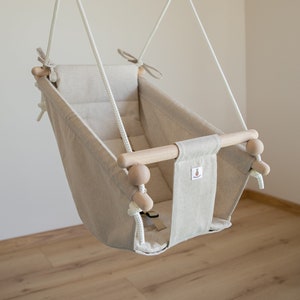 Könnte beinhalten: Eine beige Baby-Schaukel mit einer Holzstange und einem natürlichen Seil. Die Schaukel hängt von der Decke und hat einen gepolsterten Sitz.
