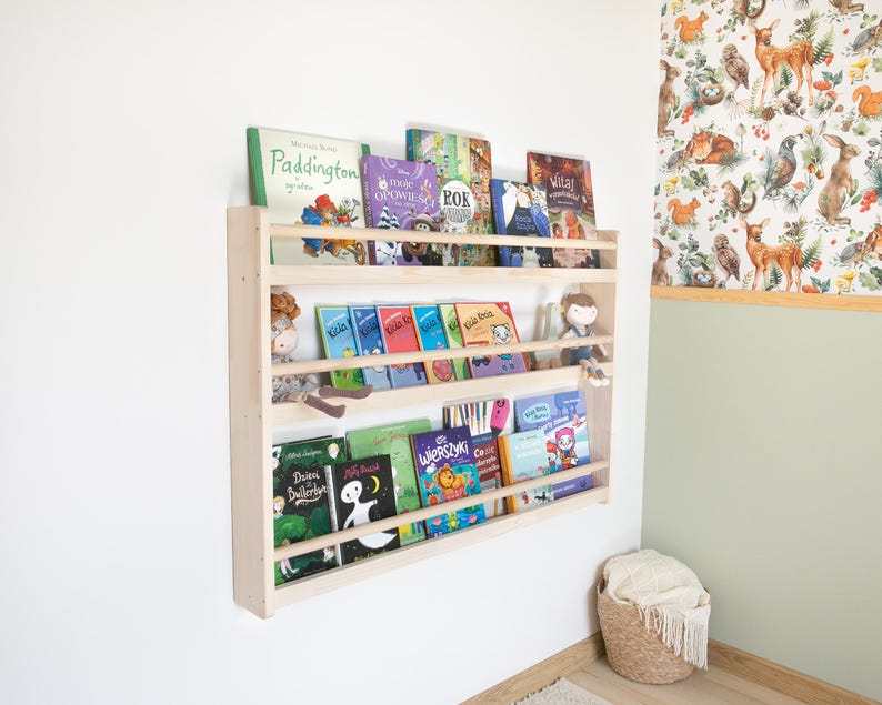 May include: A light-colored wooden wall-mounted bookshelf with three horizontal tiers, holding various children's books with colorful covers and two small fabric dolls sitting on the middle shelf, mounted on a plain white wall.