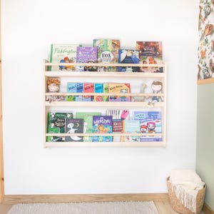 May include: A light-colored wooden wall-mounted bookshelf with three horizontal tiers. The shelves are filled with various children's books featuring colorful covers and small plush toys placed between the books on the middle shelf.