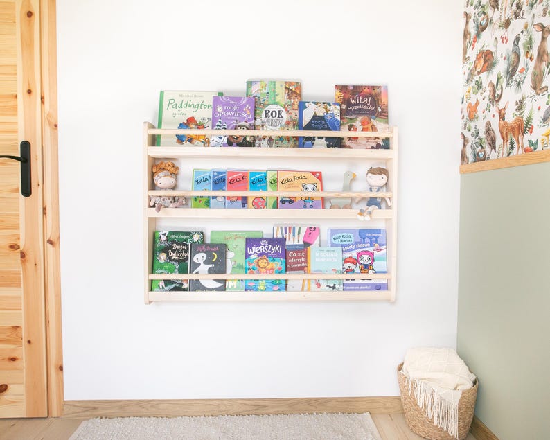 May include: A light-colored wooden wall-mounted bookshelf with three horizontal tiers. The shelves are filled with various children's books featuring colorful covers and small plush toys placed between the books on the middle shelf.