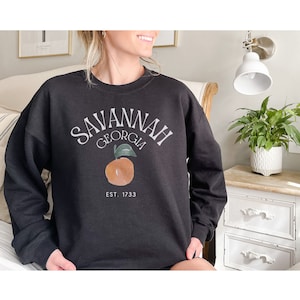 May include: Black sweatshirt with the text "SAVANNAH GEORGIA" in a curved arch, an orange graphic, and "EST. 1733" below. The sweatshirt is a crew neck style.