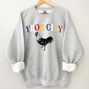 May include: A gray sweatshirt with the text "YBOR CITY" in red, yellow, blue, and white letters. A black rooster illustration is below the text.