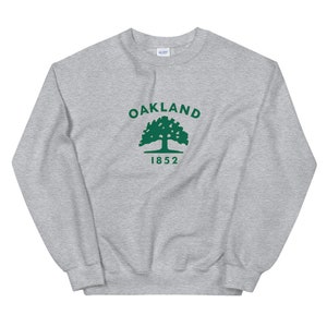 Könnte beinhalten: Ein grauer Pullover mit einem grünen Oakland-Logo, das einen Baum und das Jahr 1852 zeigt.