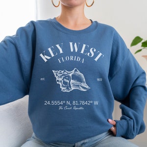 May include: A blue sweatshirt with a white graphic print that says "KEY WEST FLORIDA est. 1822" with a seashell illustration and coordinates "24.5554° N, 81.7842° W The Conch Republic".