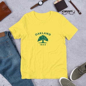 Könnte beinhalten: Gelbes T-Shirt mit kurzen Ärmeln, auf dem vorne ein grüner Baum und der Text "Oakland 1852" aufgedruckt sind.