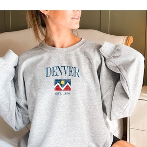 May include: A gray sweatshirt with the word "DENVER" printed in blue on the front. Below the word is a graphic of the Denver flag with the text "EST. 1858" underneath.