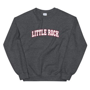 Puede incluir: Una sudadera gris oscuro con la inscripción "LITTLE ROCK" en blanco, contorneada en rojo, en el pecho. La tipografía tiene un aspecto envejecido. Una prenda casual y cómoda.