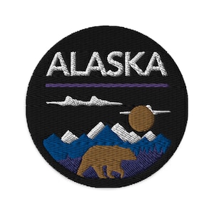 May include: A black and white embroidered patch with the word "Alaska" in white text. The patch features a brown bear walking in front of a blue and white mountain range with a brown sun in the background.